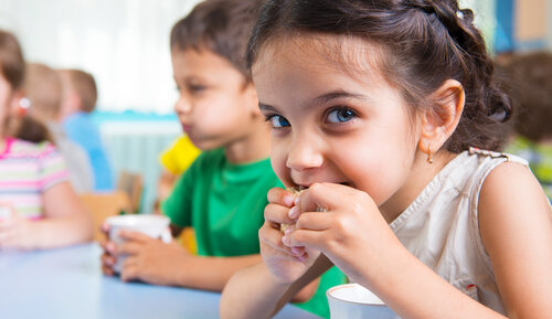 A young girl looking at the camera and eating a snack at a table with other children, illustrating coeliac disease