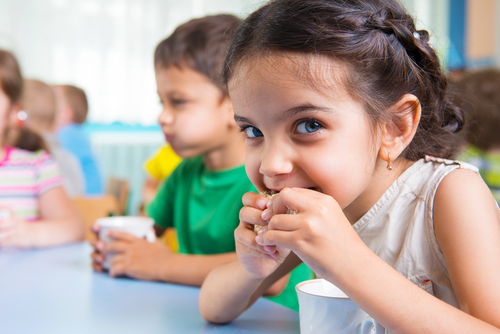 A young girl looking at the camera and eating a snack at a table with other children, illustrating coeliac disease