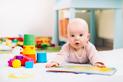 Baby lying on stomach looking at a picture book next to colorful stacking cups