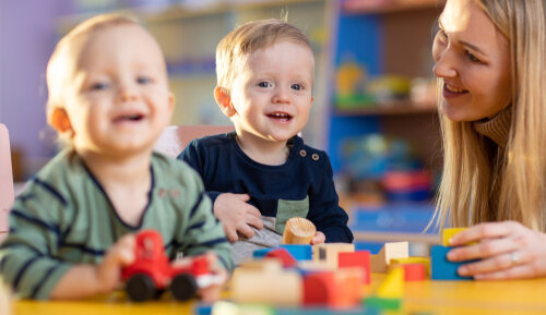 A nursery teacher smiling warmly while sitting at a table with two toddlers playing with wooden toy blocks and a red toy car.