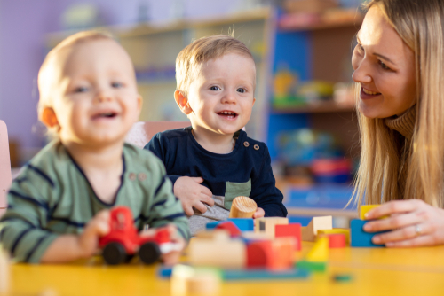 A nursery teacher smiling warmly while sitting at a table with two toddlers playing with wooden toy blocks and a red toy car.