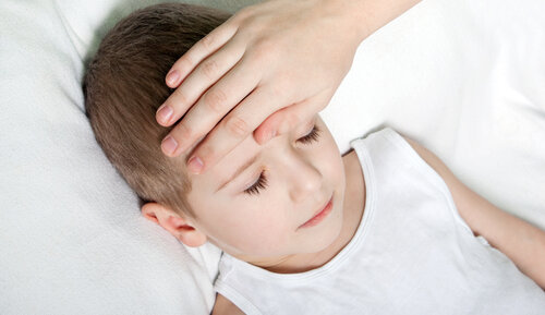 An adult's hand resting on the forehead of a young boy lying down with closed eyes.