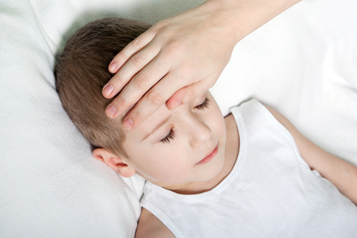 An adult's hand resting on the forehead of a young boy lying down with closed eyes.